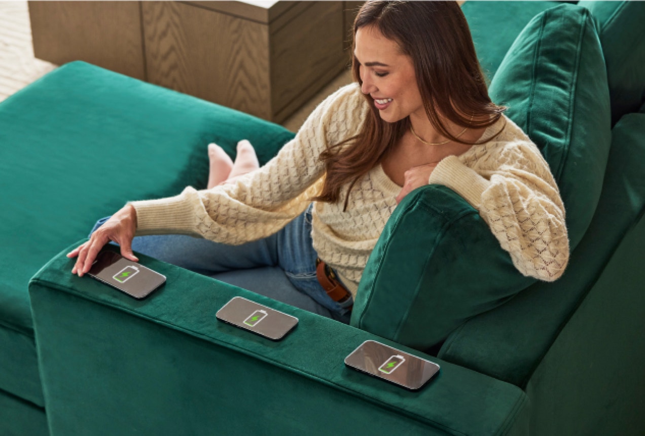 A woman is charging phones using her Lovesac Charge Side arm piece, which is built into her green sectional couch.